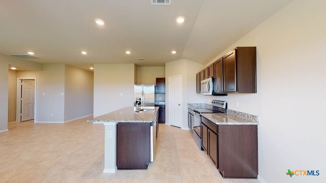 kitchen featuring stainless steel appliances, light stone countertops, a center island with sink, and dark brown cabinetry