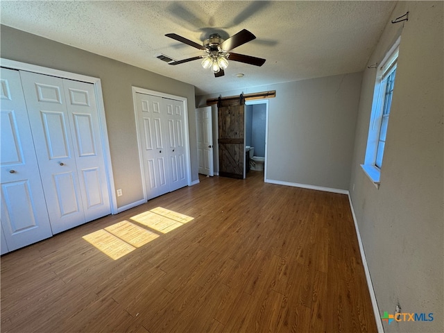 unfurnished bedroom featuring a textured ceiling, two closets, ceiling fan, wood-type flooring, and a barn door