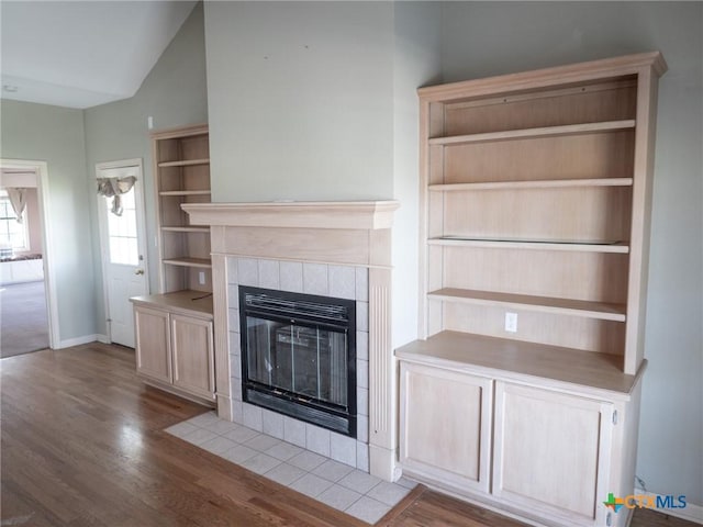 unfurnished living room featuring baseboards, wood finished floors, a tiled fireplace, and vaulted ceiling