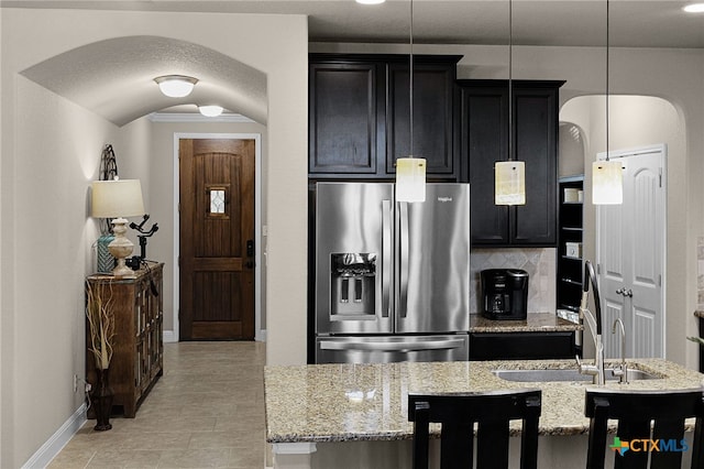 kitchen featuring a kitchen island with sink, backsplash, hanging light fixtures, light stone counters, and stainless steel fridge with ice dispenser