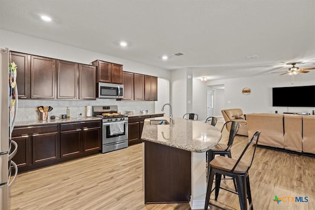 kitchen with sink, a kitchen island with sink, stainless steel appliances, light stone countertops, and light hardwood / wood-style floors