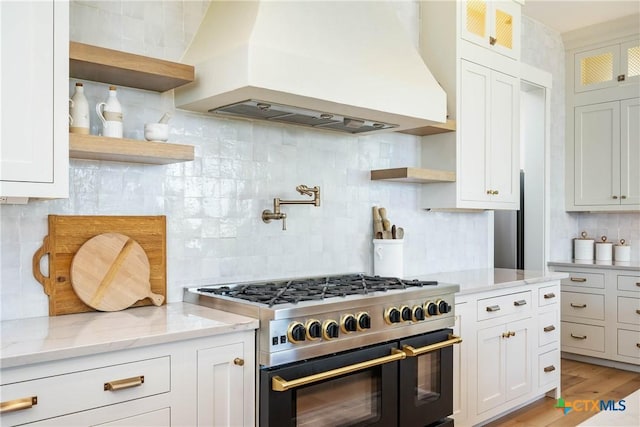 kitchen featuring custom exhaust hood, range with two ovens, open shelves, glass insert cabinets, and tasteful backsplash