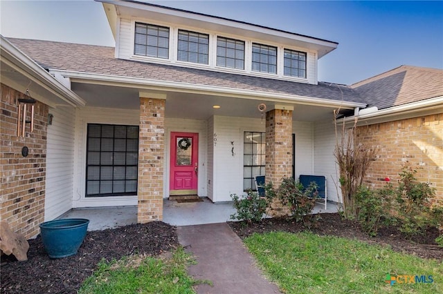 doorway to property with brick siding, covered porch, and roof with shingles