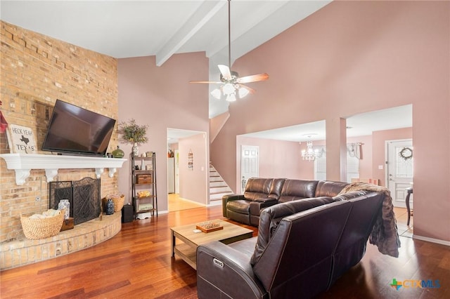 living room featuring beamed ceiling, ceiling fan with notable chandelier, wood finished floors, a brick fireplace, and stairs