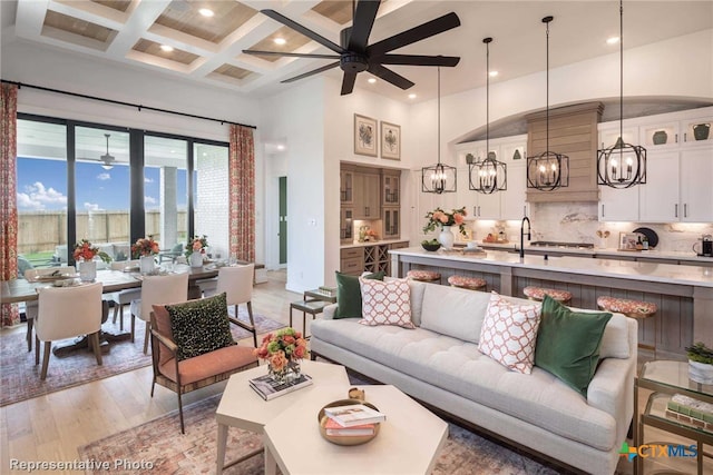 living room with coffered ceiling, light wood-type flooring, beamed ceiling, ceiling fan with notable chandelier, and a high ceiling