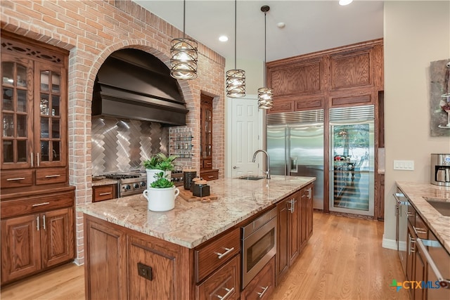 kitchen with brown cabinetry, light wood-style flooring, built in appliances, custom exhaust hood, and a sink