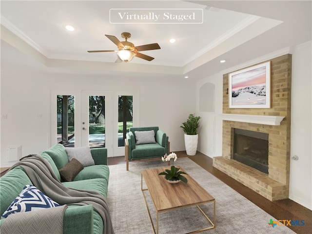 living room featuring a brick fireplace, dark hardwood / wood-style flooring, ceiling fan, crown molding, and a raised ceiling