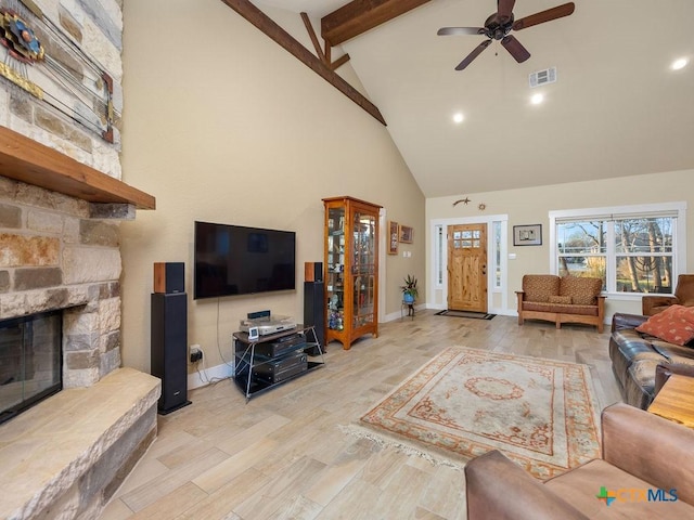 living room with visible vents, a ceiling fan, beamed ceiling, a stone fireplace, and light wood-style floors