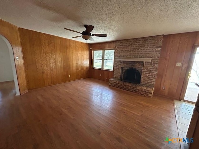 unfurnished living room with arched walkways, a ceiling fan, wood finished floors, wood walls, and a brick fireplace