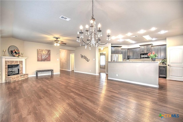 unfurnished living room with visible vents, ceiling fan with notable chandelier, dark wood-style floors, baseboards, and a brick fireplace