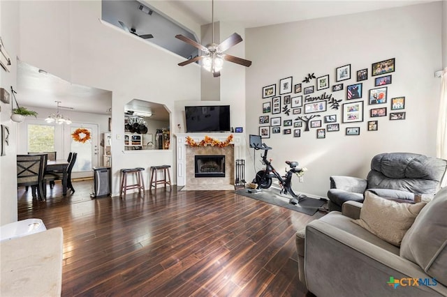 living room with ceiling fan with notable chandelier, dark wood-type flooring, a tile fireplace, and high vaulted ceiling