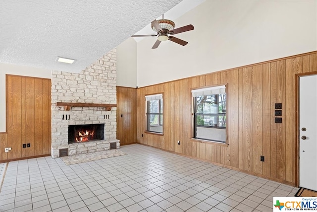 unfurnished living room featuring a stone fireplace, a textured ceiling, high vaulted ceiling, wooden walls, and ceiling fan