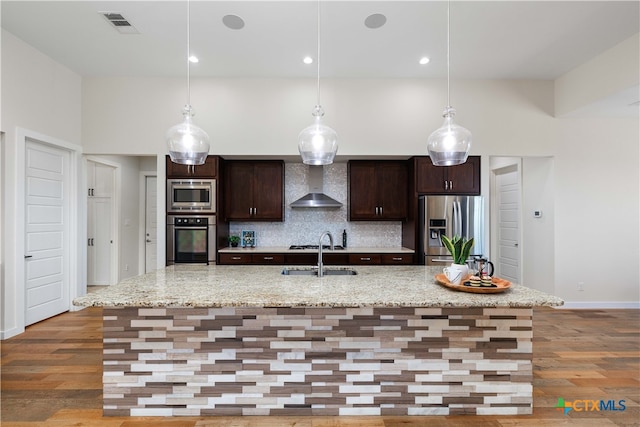 kitchen with appliances with stainless steel finishes, a center island with sink, wall chimney range hood, and light hardwood / wood-style flooring