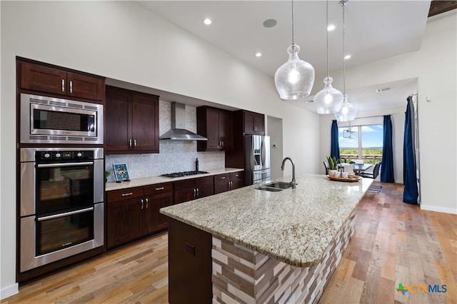 kitchen with a center island with sink, stainless steel appliances, wall chimney range hood, sink, and light hardwood / wood-style flooring