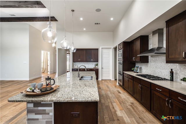 kitchen with light hardwood / wood-style floors, sink, an island with sink, stainless steel gas stovetop, and wall chimney range hood