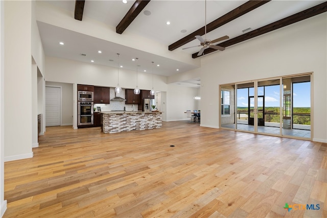 unfurnished living room featuring ceiling fan, a towering ceiling, beamed ceiling, and light wood-type flooring