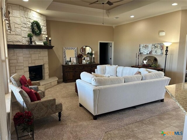 carpeted living room with ceiling fan, a stone fireplace, a raised ceiling, and ornamental molding