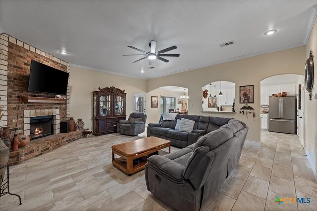 living area with ceiling fan, visible vents, arched walkways, a brick fireplace, and ornamental molding