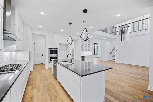 kitchen with wall chimney range hood, white cabinetry, stainless steel appliances, a center island with sink, and decorative light fixtures