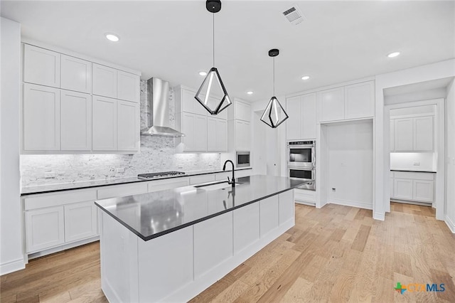 kitchen featuring wall chimney exhaust hood, white cabinetry, decorative light fixtures, stainless steel appliances, and a kitchen island with sink