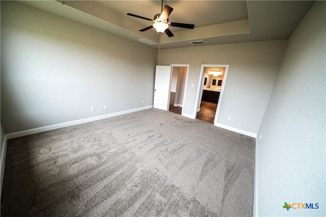 unfurnished bedroom featuring a raised ceiling, dark carpet, ceiling fan, and ensuite bath