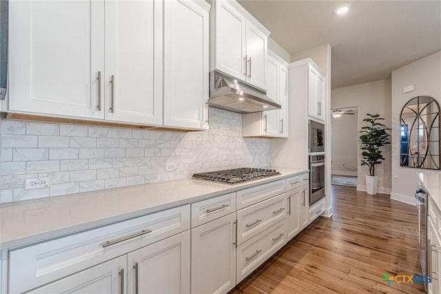 kitchen with under cabinet range hood, tasteful backsplash, white cabinetry, stainless steel appliances, and light wood finished floors