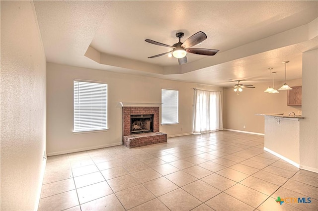 unfurnished living room featuring a brick fireplace, ceiling fan, a raised ceiling, a textured ceiling, and light tile patterned floors