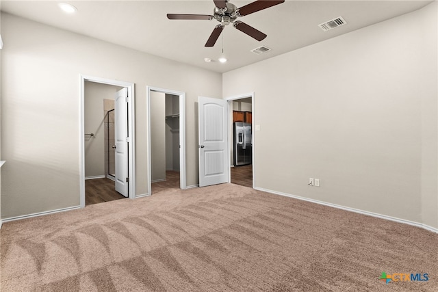 unfurnished bedroom with a walk in closet, ensuite bath, stainless steel refrigerator with ice dispenser, dark colored carpet, and ceiling fan