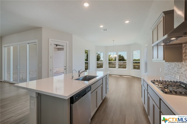 kitchen featuring sink, an island with sink, gray cabinets, wall chimney range hood, and appliances with stainless steel finishes