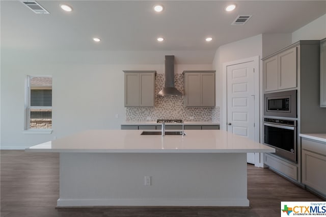 kitchen with appliances with stainless steel finishes, wall chimney exhaust hood, dark wood-type flooring, and a center island with sink