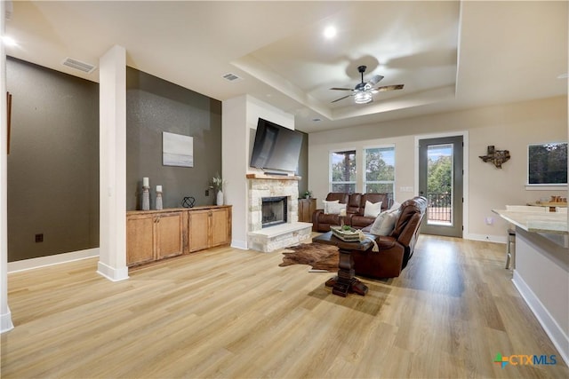 living room with a tray ceiling, a stone fireplace, ceiling fan, and light hardwood / wood-style floors