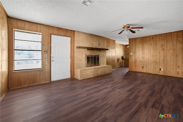 unfurnished living room with wood walls, dark wood-type flooring, a brick fireplace, ceiling fan, and a textured ceiling