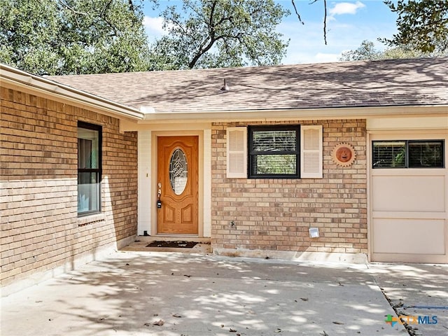 entrance to property featuring brick siding, a garage, driveway, and roof with shingles