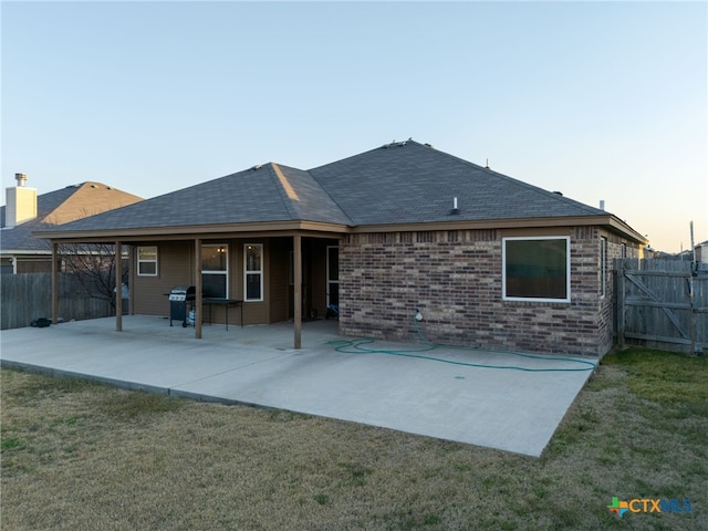 back of house at dusk featuring brick siding, a shingled roof, a lawn, a patio area, and fence