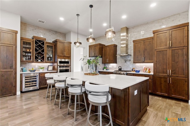 kitchen with beverage cooler, wall chimney exhaust hood, hanging light fixtures, a spacious island, and appliances with stainless steel finishes