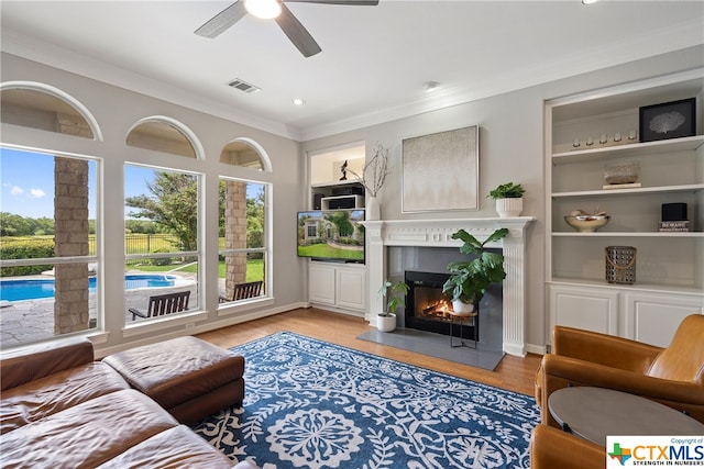 sitting room with built in shelves, light wood-type flooring, a glass covered fireplace, and crown molding