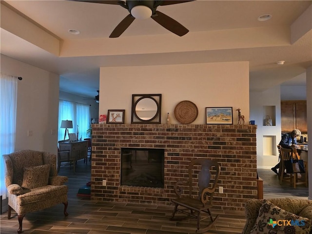 living room with a brick fireplace, a tray ceiling, dark wood-type flooring, and ceiling fan