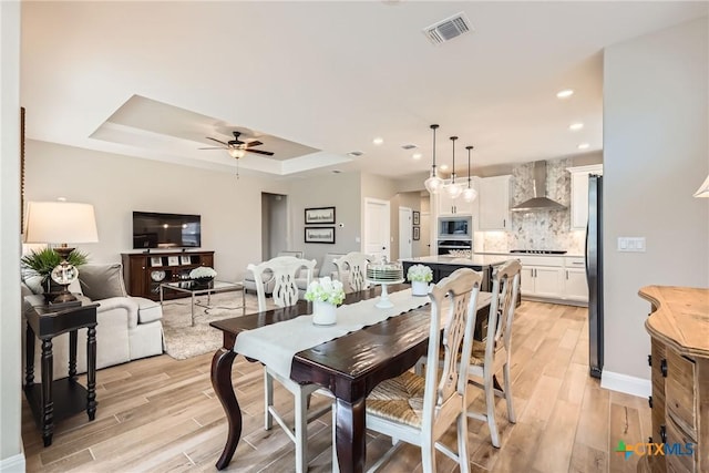 dining space with light wood-type flooring, a tray ceiling, visible vents, and a ceiling fan