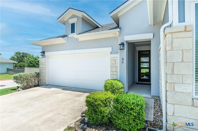 view of exterior entry featuring roof with shingles, stucco siding, concrete driveway, fence, and stone siding