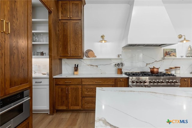 kitchen featuring premium range hood, appliances with stainless steel finishes, tasteful backsplash, light stone counters, and light hardwood / wood-style floors