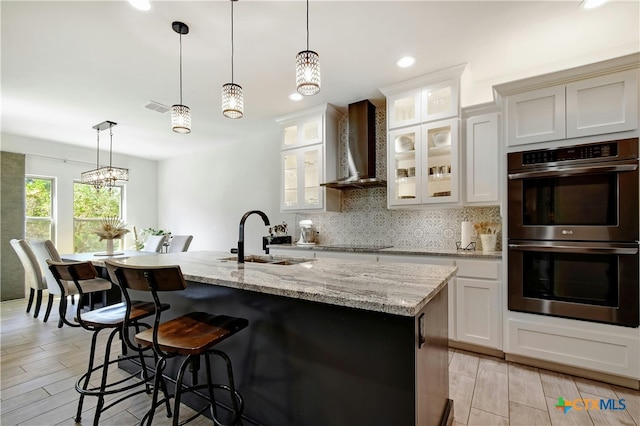 kitchen featuring a center island with sink, white cabinetry, wall chimney exhaust hood, and double oven