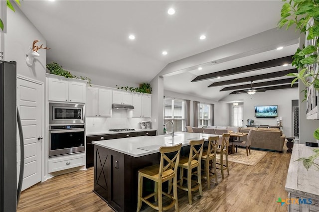 kitchen featuring vaulted ceiling with beams, white cabinets, open floor plan, appliances with stainless steel finishes, and an island with sink