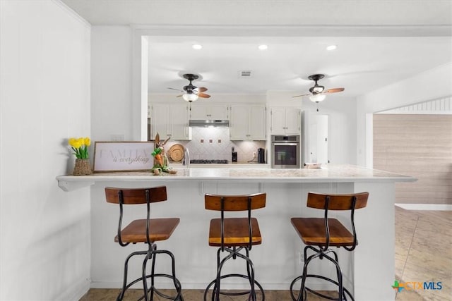 kitchen featuring ceiling fan, under cabinet range hood, a kitchen bar, decorative backsplash, and a peninsula