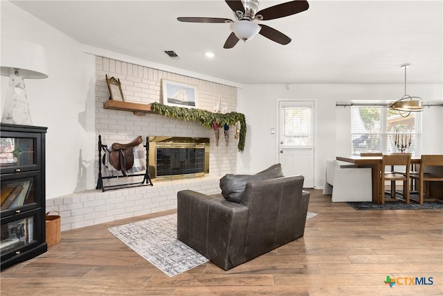 living room with wood-type flooring, ceiling fan with notable chandelier, and a brick fireplace