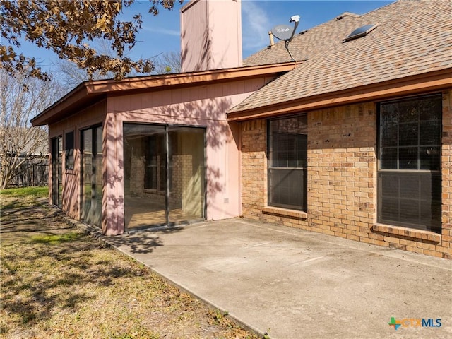 exterior space featuring brick siding, roof with shingles, a chimney, a patio area, and fence