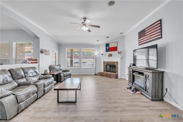 living room with baseboards, ceiling fan, ornamental molding, a brick fireplace, and light wood-type flooring