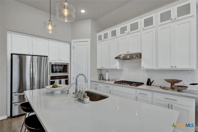 kitchen featuring a sink, under cabinet range hood, appliances with stainless steel finishes, glass insert cabinets, and hanging light fixtures