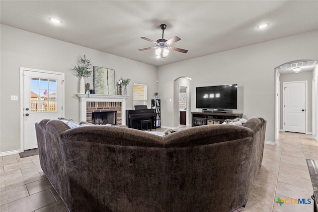 living room featuring light tile patterned floors, arched walkways, a brick fireplace, and a ceiling fan