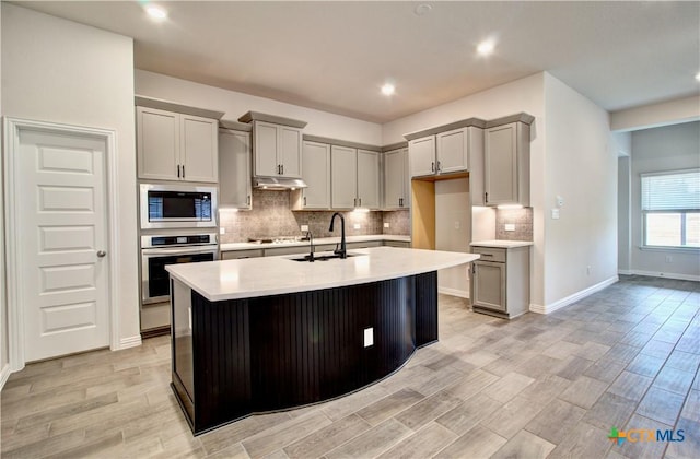 kitchen with stainless steel oven, a kitchen island with sink, gray cabinets, and built in microwave