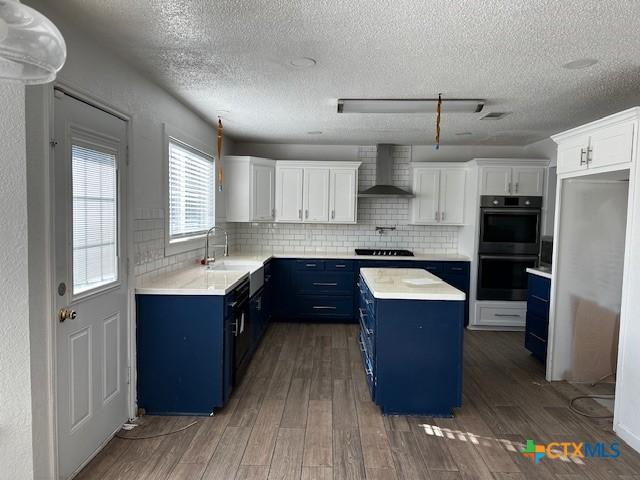 kitchen with black appliances, blue cabinets, wall chimney exhaust hood, a kitchen island, and white cabinetry